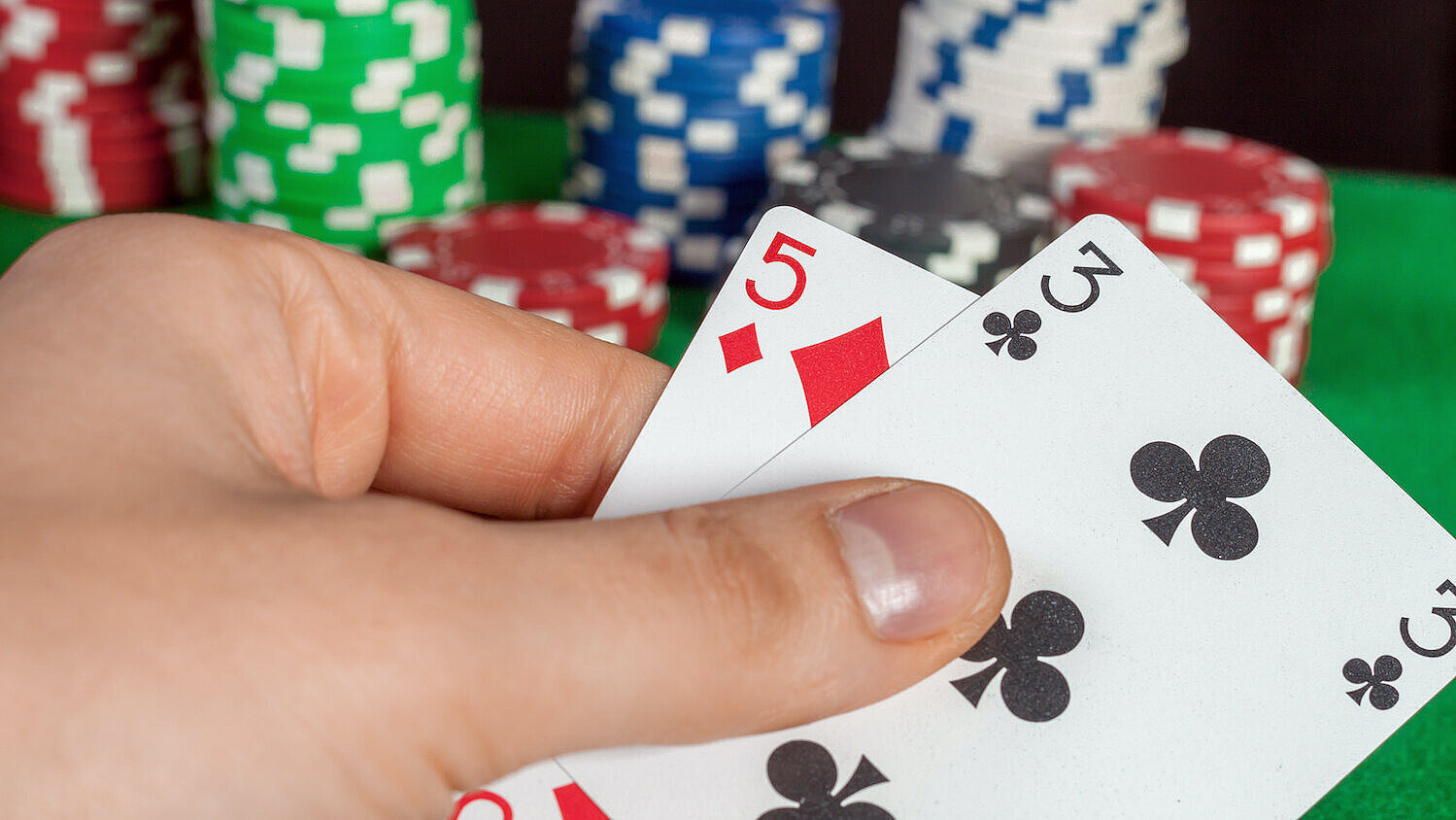 Eine Person hält die Spielkarten Karo 5 und Kreuz 3 in der Hand, im Hintergrund liegen Pokerchips. 