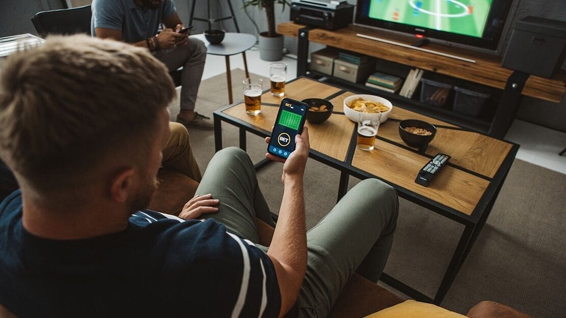 Mann sitzt auf dem Sofa, schaut Fußball im Fernsehen und hält ein Smartphone mit Wett-App in der Hand