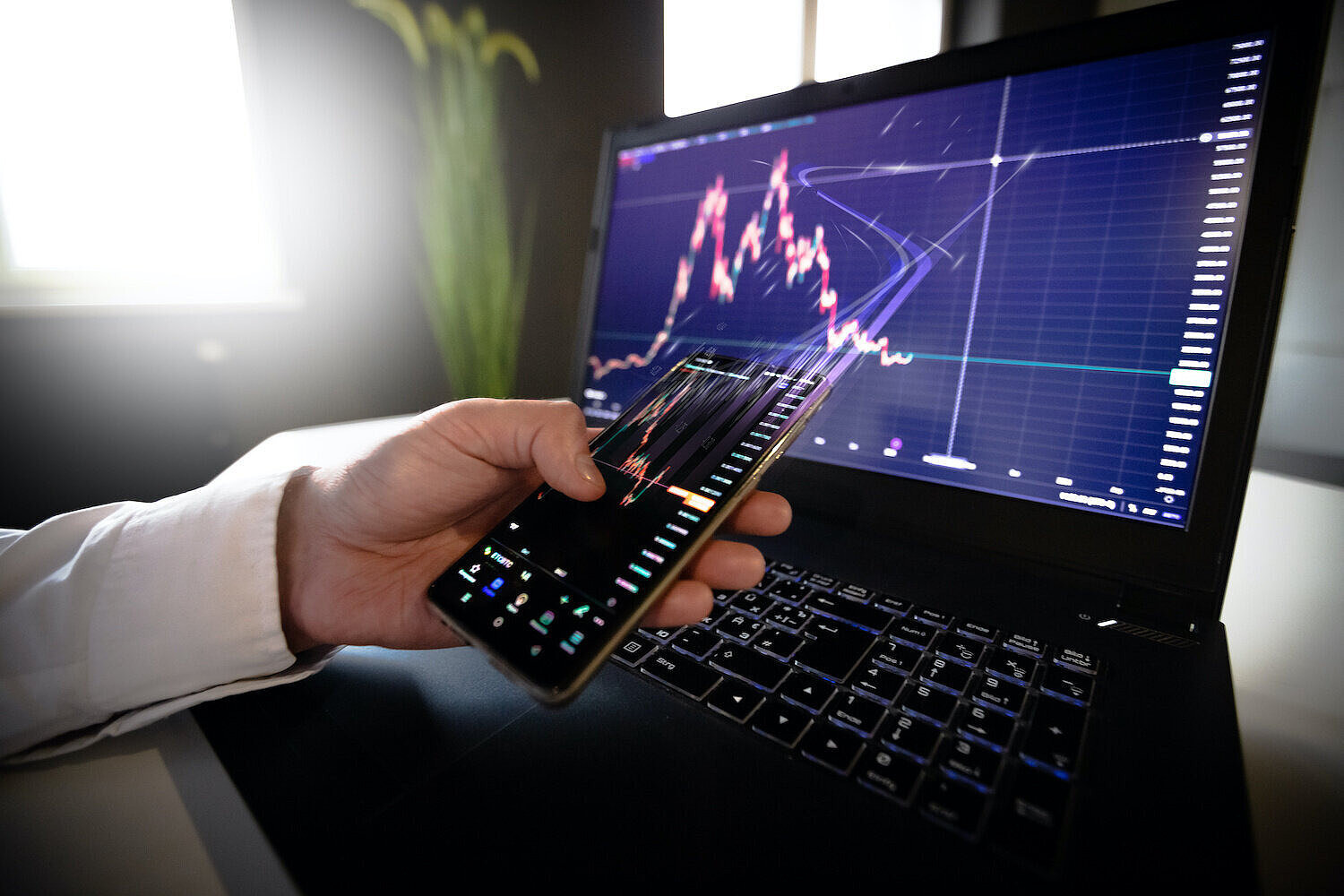 Person hat Handy in der Hand und Laptop vor sich und auf beiden Bildschirmen befinden sich Stockcharts mit Aktienkursen 