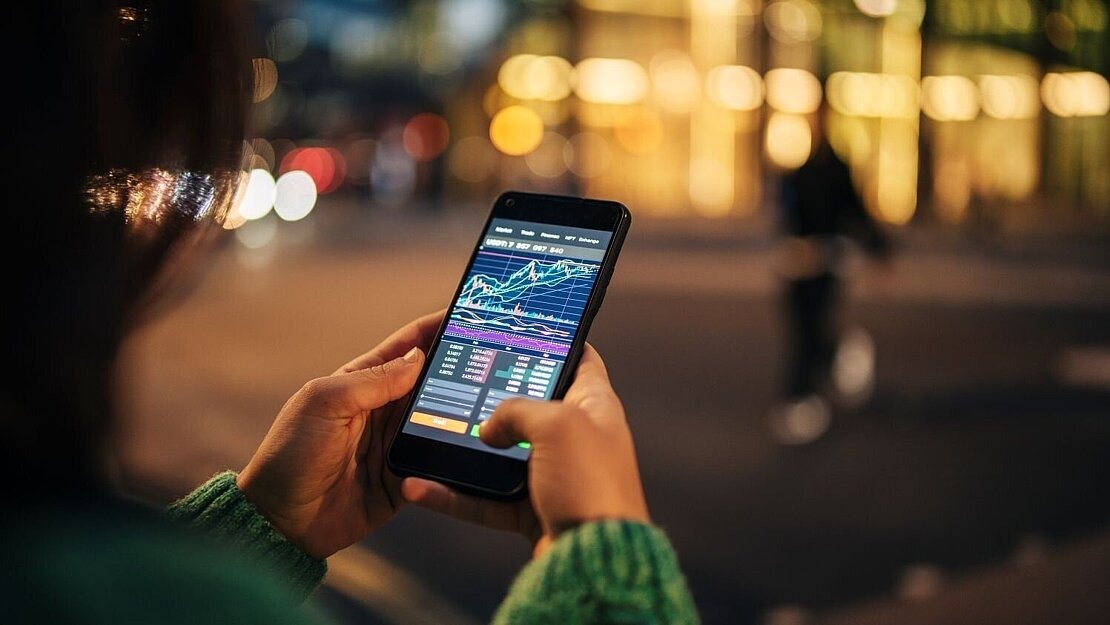 Person hält abends draußen ein Smartphone mit Trading-App und Kursdiagramm in der Hand.