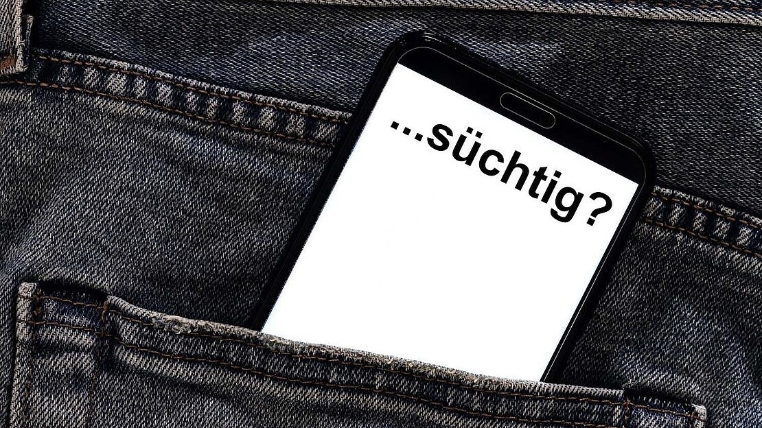 Aus einer Hosentasche ragt ein Handy auf dem "...süchtig?" steht. 