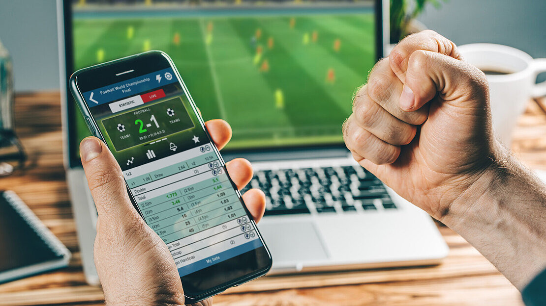 Ein Mann hält ein Smartphone in der Hand, auf dem eine Live-Sportwetten-App geöffnet ist. Die App zeigt den Spielstand eines Fußballspiels mit 2:1 an sowie verschiedene Wettquoten und Optionen. Im Hintergrund läuft dasselbe Spiel auf einem Laptop-Bildschirm.