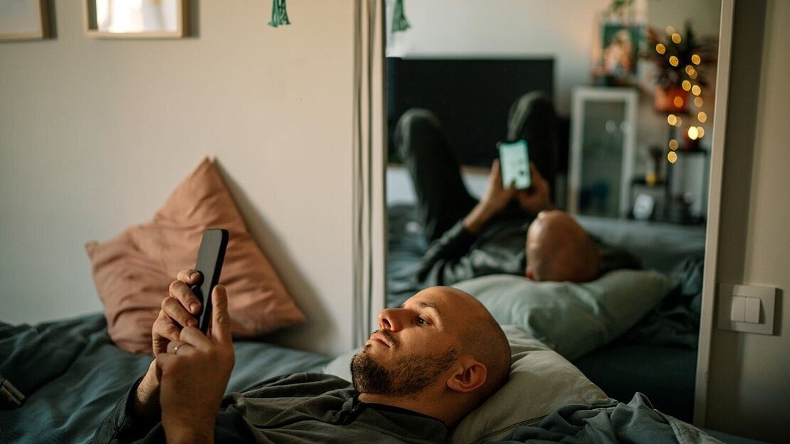 Ein Mann liegt mit Smartphone in der Hand auf dem Bett. 