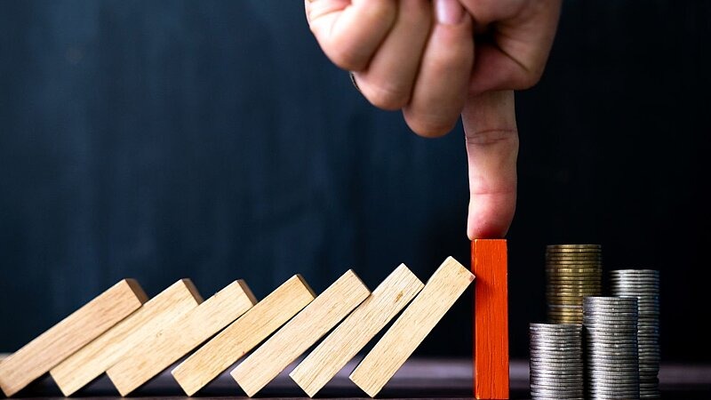 Eine Hand stoppt mit dem Finger eine Reihe umfallender Holzklötze, bevor sie einen Stapel Münzen erreicht – symbolisch für das Setzen von Grenzen und den Schutz vor finanziellen Folgen., Bild: krisanapong detraphiphat / gettyimages