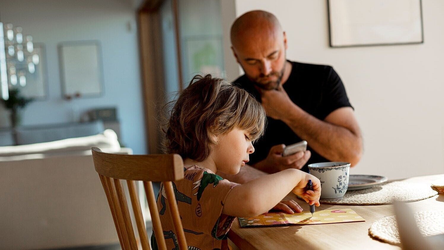 Kleines Kind sitzt am Tisch und malt, während ein Elternteil im Hintergrund auf das Handy schaut