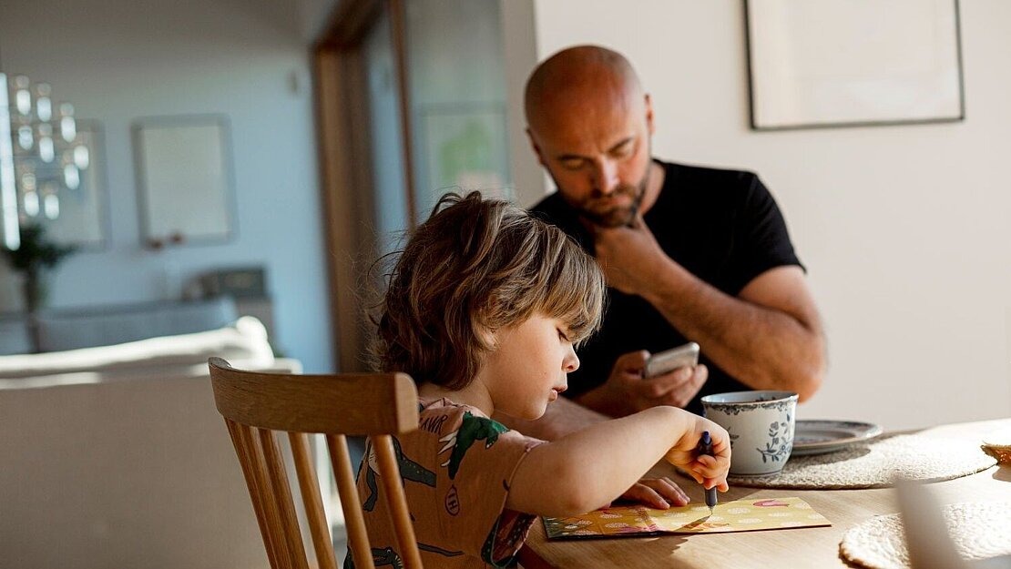 Kleines Kind sitzt am Tisch und malt, während ein Elternteil im Hintergrund auf das Handy schaut